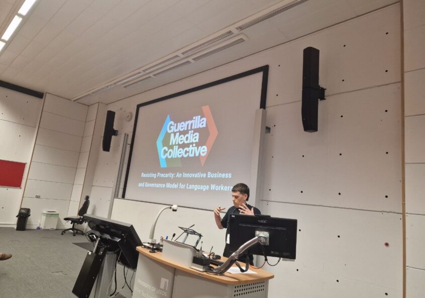 Person presenting in a university lecture theatre, with the Guerrilla Media Collective logo on the screen